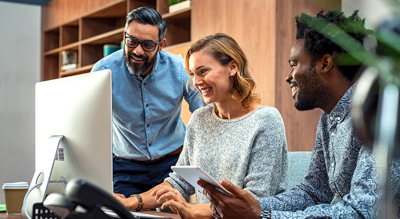 A photo of three professionally-dressed people in a wood-accented office grouped around a monitor, smiling and talking