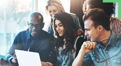 A diverse group of people gathered around a laptop, engaged in discussion and looking at the screen together.