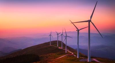 A photo of a row of wind turbines on a mountaintop against a backdrop of hazy purple mountains and a yellow and pink sunset