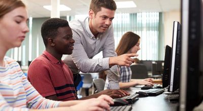 Três jovens estudantes sentados em um laboratório de informática enquanto um adulto se inclina sobre eles e aponta para uma das telas do computador